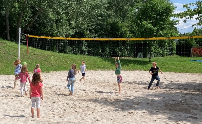 Kinder spielen gemeinsam Ball auf einem Sandplatz mit Netz.